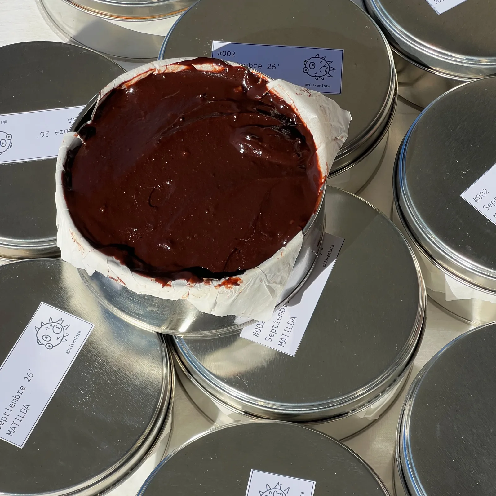Lata abierta con la torta Matilda cubierta de ganache brillante de chocolate, rodeada de latas cerradas con su etiqueta