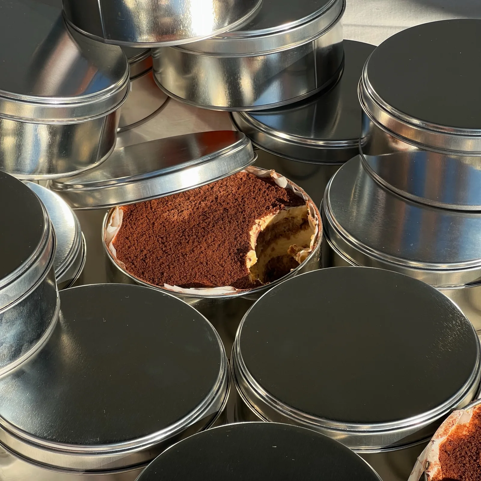 Lata abierta con tiramisú espolvoreado con cacao, entre latas cerradas de tapa negra
